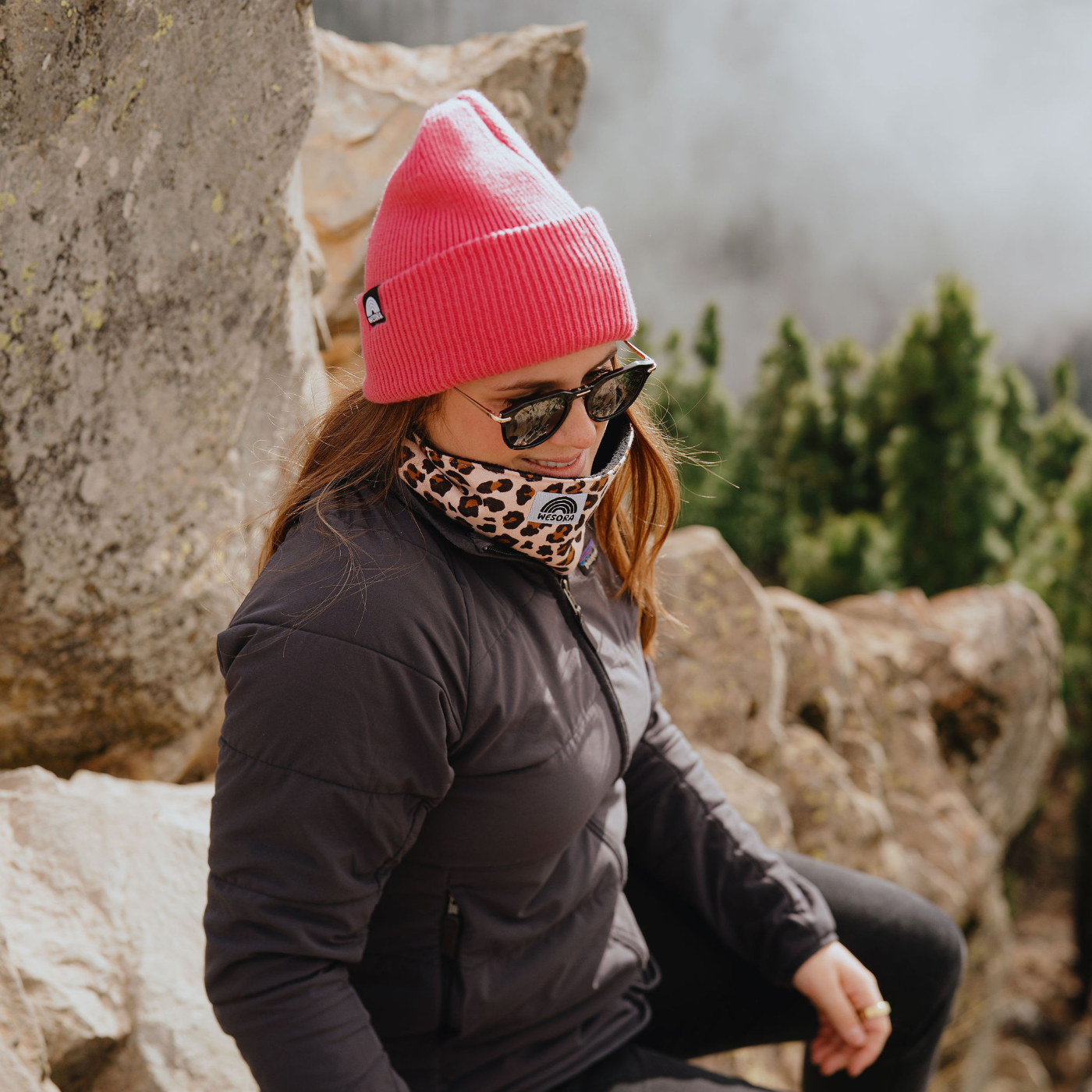 Beanie - True Pink mit doppellagigem Strickdesign, getragen von einer Person beim Wandern auf Felsen, im Hintergrund Bäume und Steinformationen.