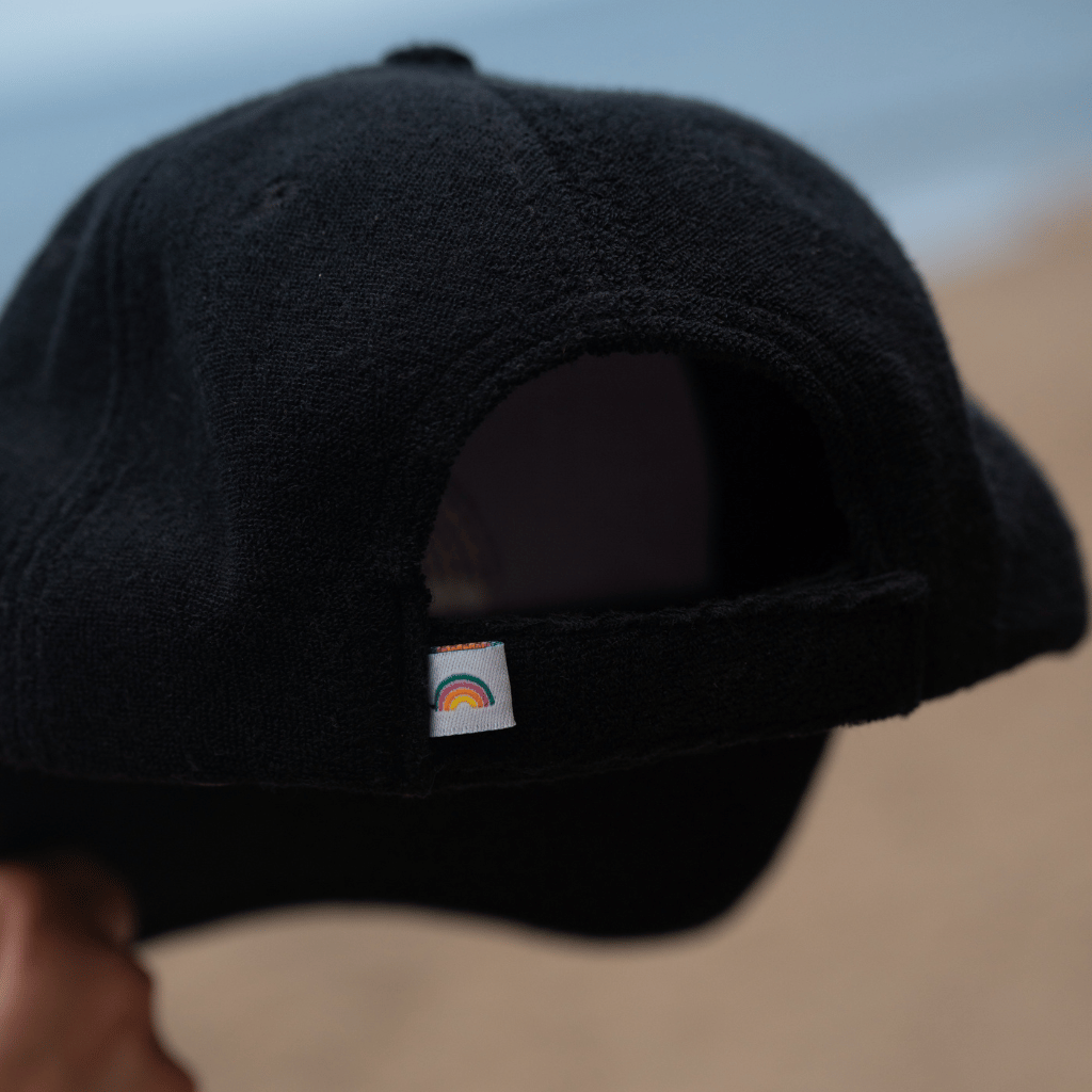Frottee Cap - All Black mit Regenbogenlogo, gehalten von einer Person, fotografiert aus der Nähe, fokussiert auf den Klettverschluss und das Label.