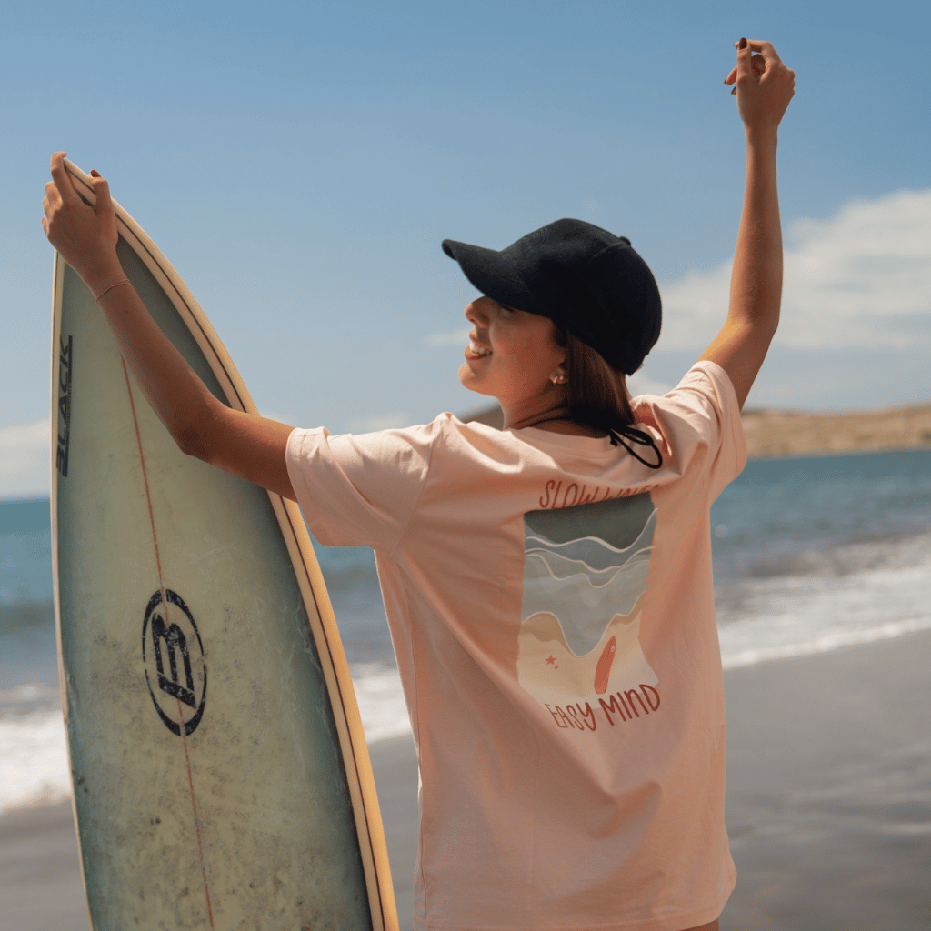 Frottee Cap - All Black mit minimalistischer Stickerei, getragen von einer Frau am Strand, die ein Surfbrett hält, in natürlicher Outdoor-Szenerie.