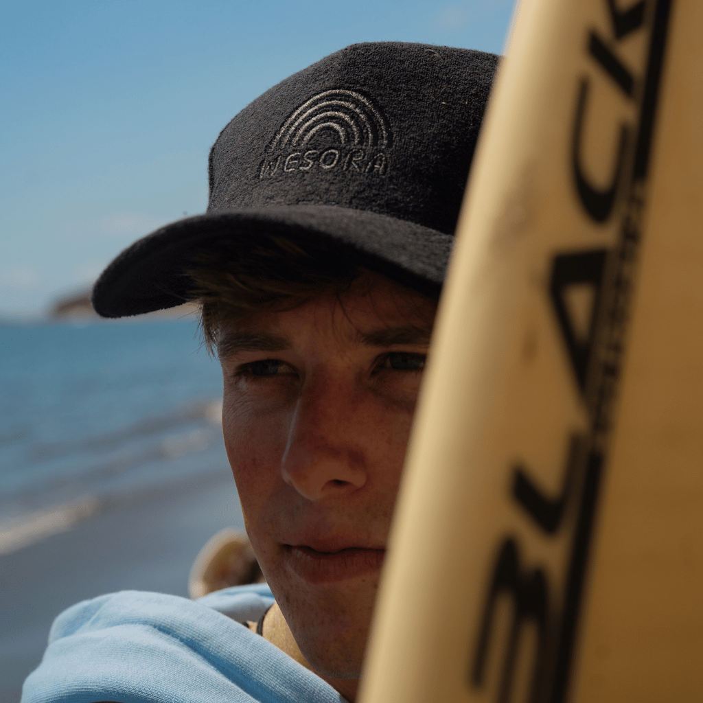 Frottee Cap - All Black mit minimalistischer Stickerei, getragen von einem Mann am Meer mit Surfboard in der Hand, Nahaufnahme der Cap, von vorne fotografiert.