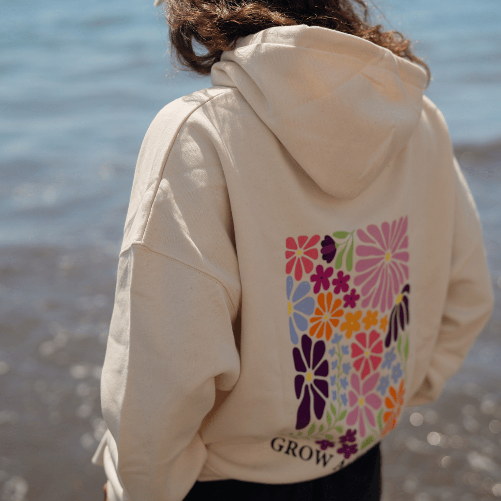Grow as you go Flower - Pullover in weiß mit farbenfrohem Blumen-Print auf dem Rücken, getragen von Person, stehend vor dem Meer, fotografiert am Strand, Nahaufnahme des Rückens.