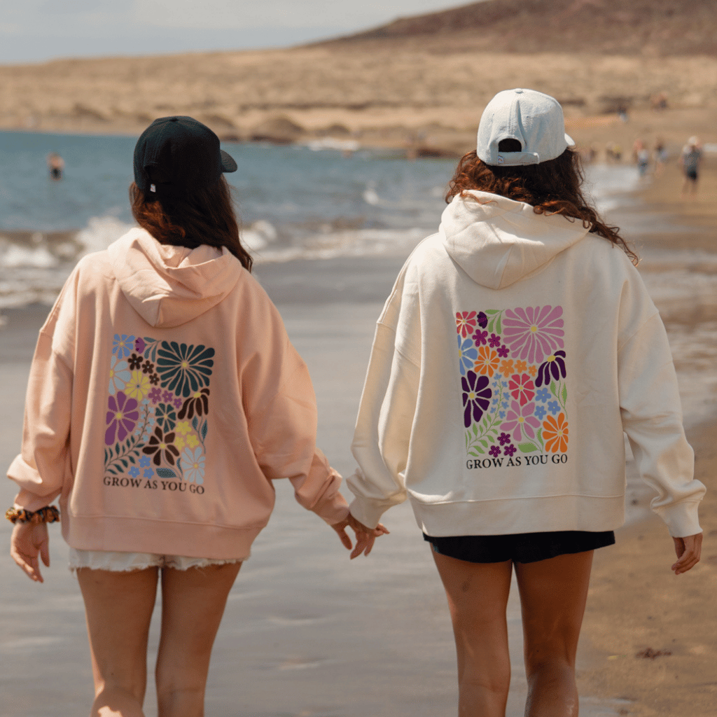 Zwei Frauen händchenhaltend und spazierend am Strand mit Blick aufs Meer, tragen den Grow as you go Flower - Pullover mit großem Blumen-Print auf dem Rücken in zwei unterschiedlichen Farben.