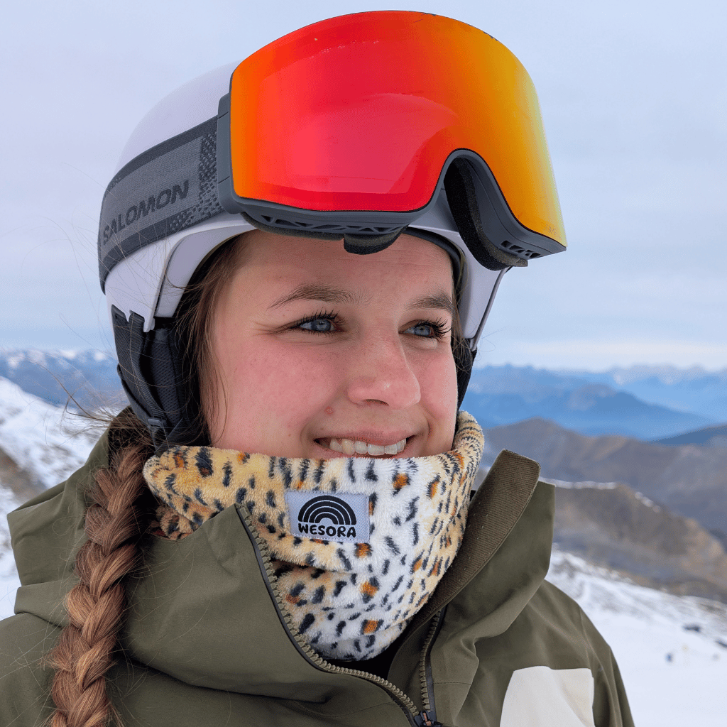 Printed Neckwarmer - Fluffy Wild Leo mit Leopardenmuster, getragen von einer Frau in Winterkleidung, lächelnd auf schneebedecktem Berghang.