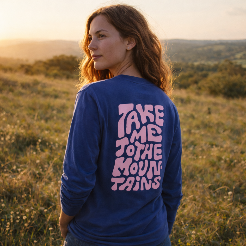 Take me to the Mountains - Unisex Longsleeve mit großem Rückenprint, getragen von junger Frau in Feldlandschaft bei Sonnenuntergang, seitliche Ansicht.