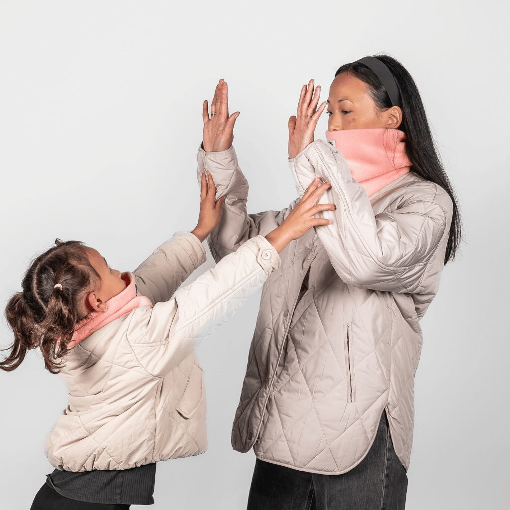 Eltern + Kind Bundle - Fleece Neckwarmer Peachy Rose, getragen von Mutter und Tochter in einem spielerischen Moment, beide mit beige Steppjacken, Hände zum High-Five erhoben.