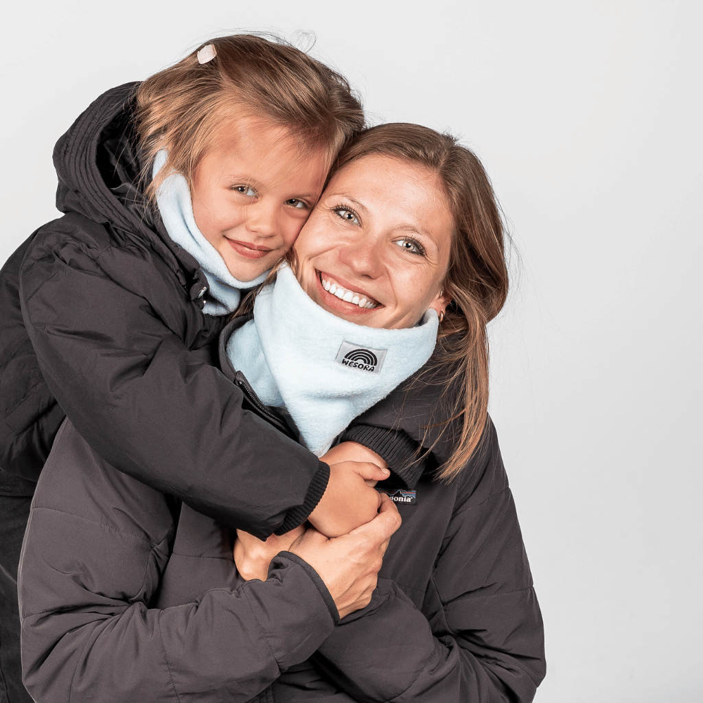 Eltern + Kind Bundle - Fleece Neckwarmer Baby Blue, getragen von lachender Frau, umarmt von Mädchen in dunklen Winterjacken, vor neutralem Hintergrund, warm und liebevoll.