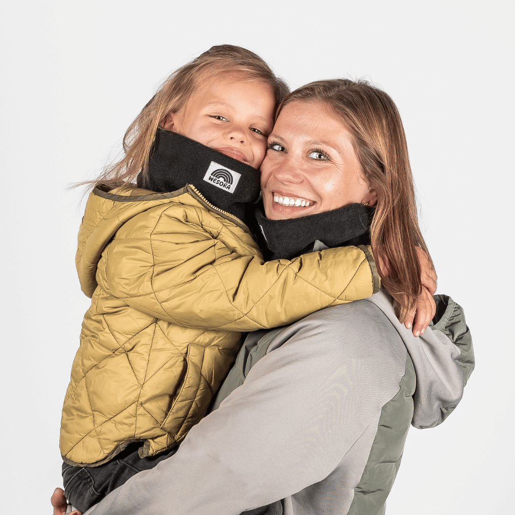 Eltern + Kind Bundle - Fleece Neckwarmer All Black, getragen von Mutter und Kind in Umarmung, fotografiert vor hellem Hintergrund, beide lächeln in die Kamera.