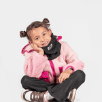 Fleece Neckwarmer -All Black Kid, getragen von einem lächelnden Kind im fluffigen Outfit, sitzt mit gekreuzten Beinen und ruht den Kopf auf der Hand.