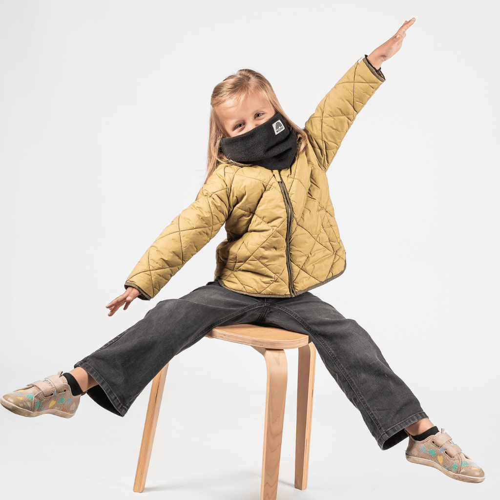 Fleece Neckwarmer -All Black Kid, getragen von einem spielenden Kind auf einem Holzstuhl, mit ausgestreckten Armen, spielerisch und dynamisch in Bewegung.