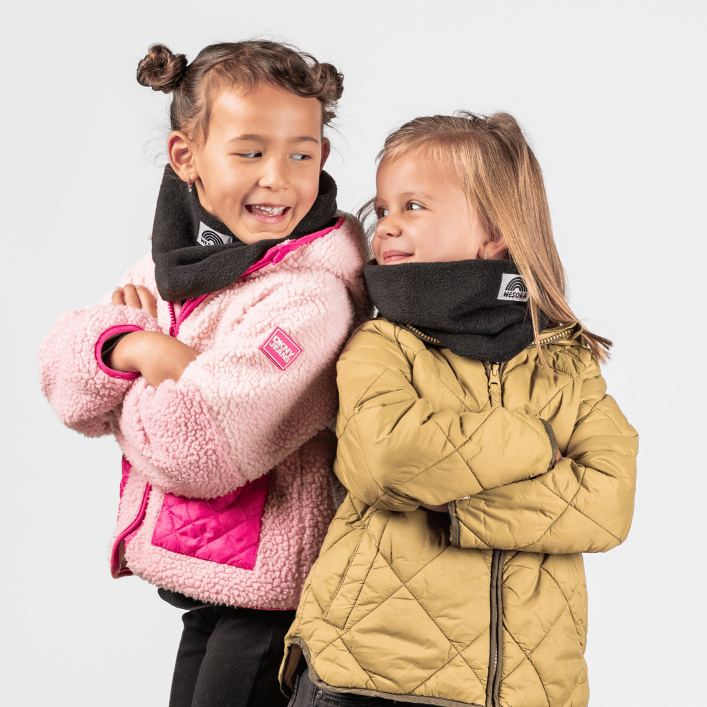 Fleece Neckwarmer -All Black Kid, getragen von zwei Kindern in warmen Jacken, die sich fröhlich anschauen, fotografiert in einem spielerischen Moment.