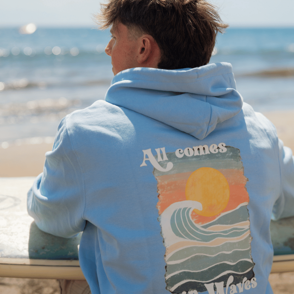 All Comes in Waves - Unisex T-Shirt mit großem Rückenprint, getragen von einem Mann auf einer Bank mit Surfbrett, Nahaufnahme zeigt Detailansicht.