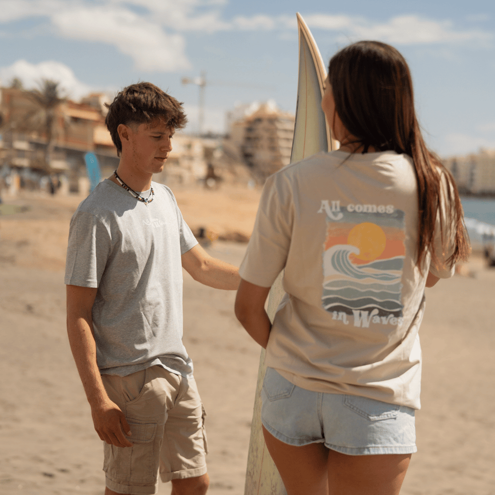 All comes in Waves - Unisex Tshirt mit großem Rückenprint, getragen von einem Mann am Strand, neben einer Frau mit Surfbrett, Nahaufnahme von Shorts.