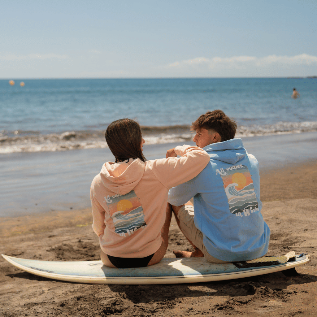 All comes in Waves - Unisex Pullover mit maritimem Design, getragen von Paar auf Surfbrett am Strand, im entspannten Outdoor-Setting.