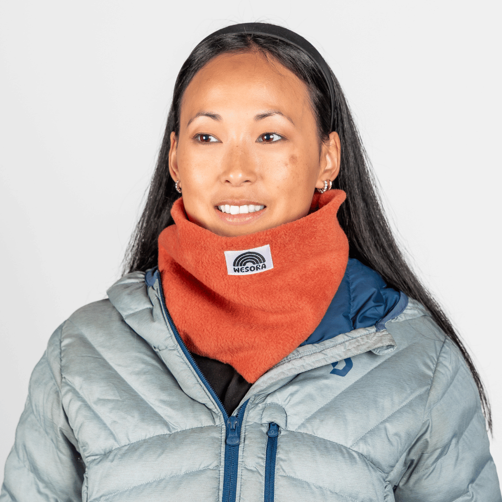 Fleece Neckwarmer - Autumn Nectar, in leuchtendem Orange, getragen von einer Frau mit dunklen Haaren, fotografiert vor hellem Hintergrund.