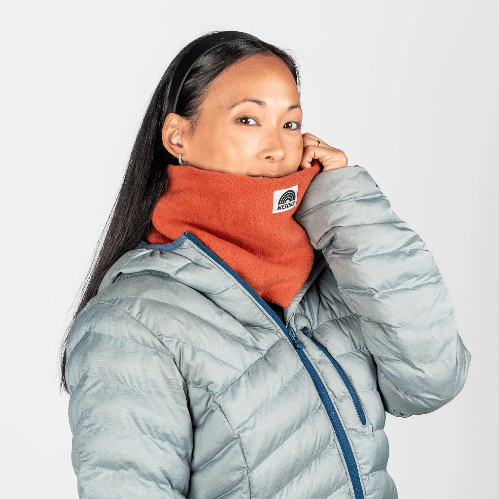 Fleece Neckwarmer - Autumn Nectar in leuchtendem Orange mit Regenbogen-Logo, getragen von Person mit grauer Jacke, vor hellem Hintergrund.