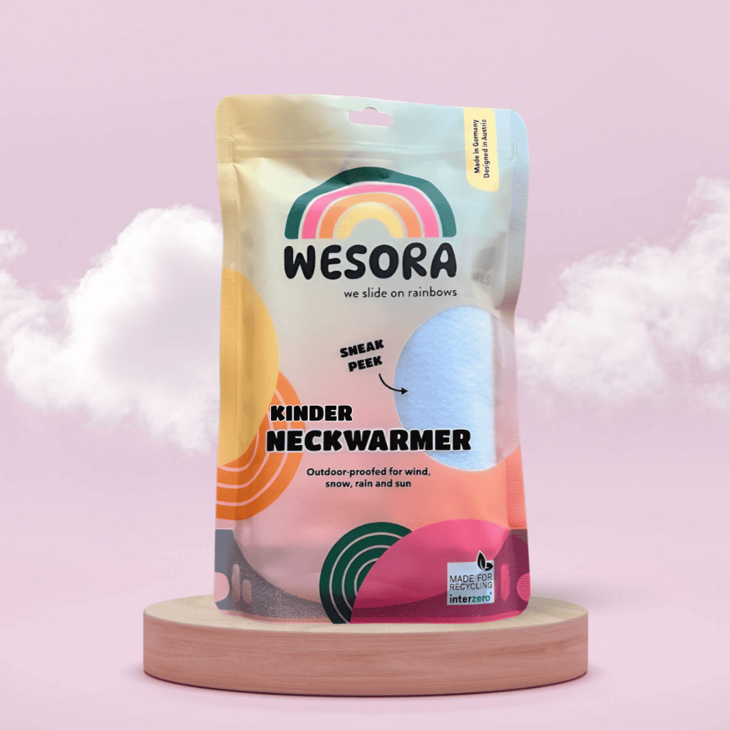 Fleece Neckwarmer - Candy Rose Kid in bunter Standbeutel-Verpackung mit Sichtfenster, präsentiert auf Holzpodest vor rosa Wolkenhintergrund. Ideal für winterliche Abenteuer.