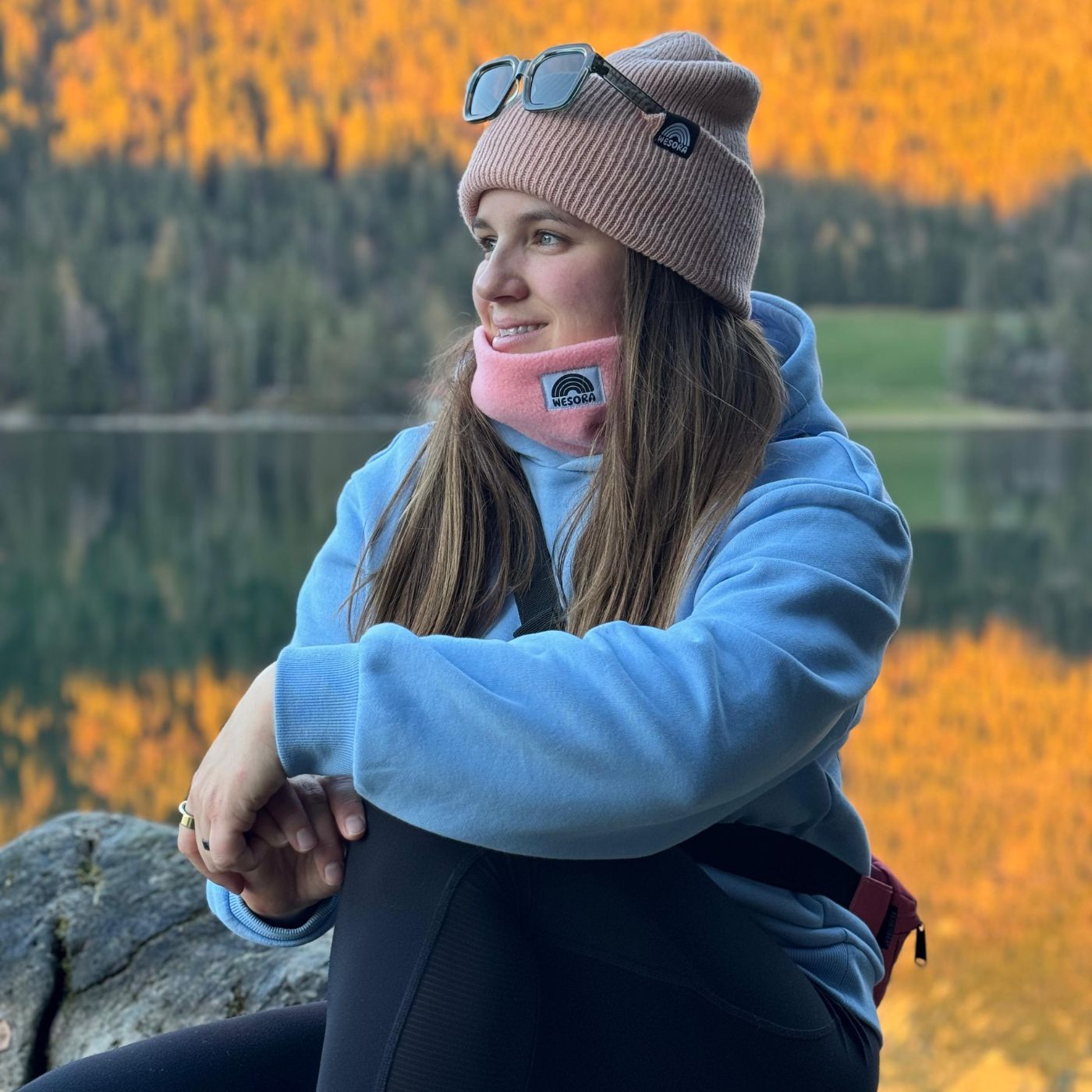 Beanie - Peach Fuzz mit doppellagigem Strickdesign, getragen von einer Frau am See bei Sonnenuntergang, sitzt auf einem Felsen.