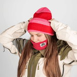 Beanie - Pink Berry Twist mit gestreiftem Design, getragen von Person mit langem Haar, passend zum roten Fleece-Nackenwärmer und zweifarbiger Jacke.