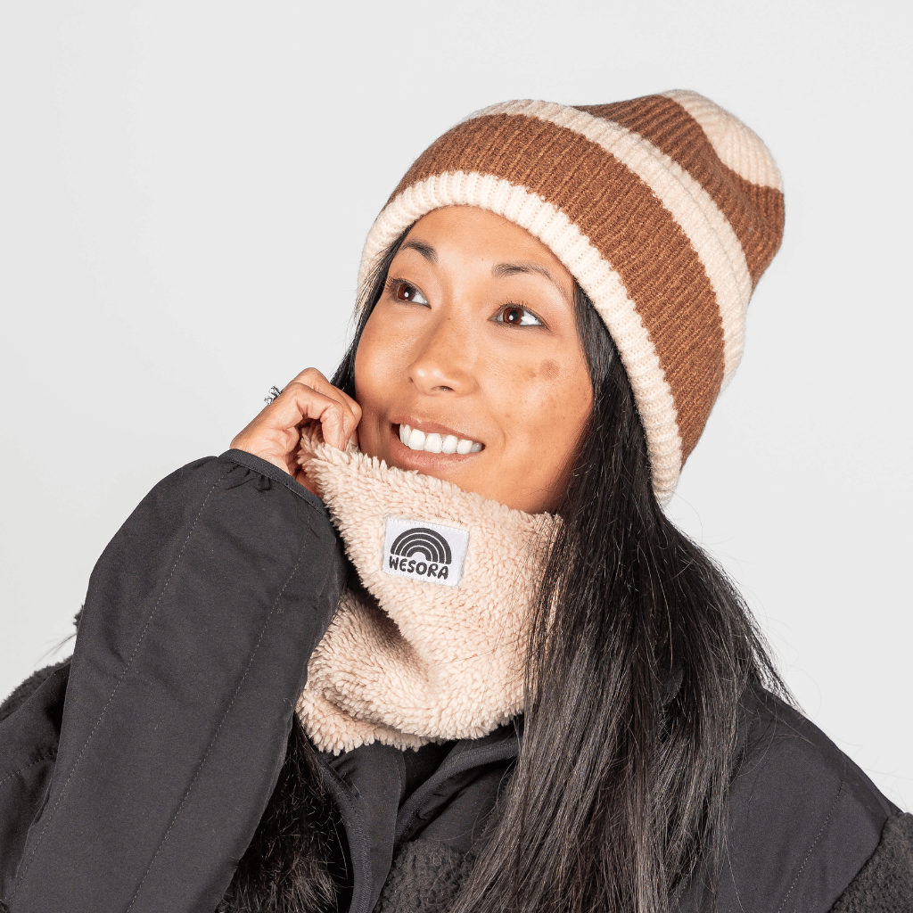 Beanie - Toffee Twist mit braun-cremem Streifen, getragen von einer lächelnden Person, kombiniert mit schwarzem Jacke und beigem, sherpa-style Neck Gaiter.