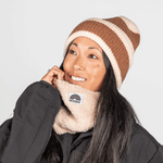 Beanie - Toffee Twist mit braun-cremem Streifen, getragen von einer lächelnden Person, kombiniert mit schwarzem Jacke und beigem, sherpa-style Neck Gaiter.