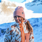 Beanie - Peach Fuzz mit geripptem Design, getragen von junger Frau in winterlicher Bergkulisse, lächelnd vor schneebedeckten Gipfeln und klarem Himmel.