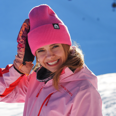 Beanie - True Pink mit doppellagigem Strickdesign, getragen von lächelnder Person in winterlicher Landschaft, Hand am Hut, fotografiert in sonniger Schneeszene.