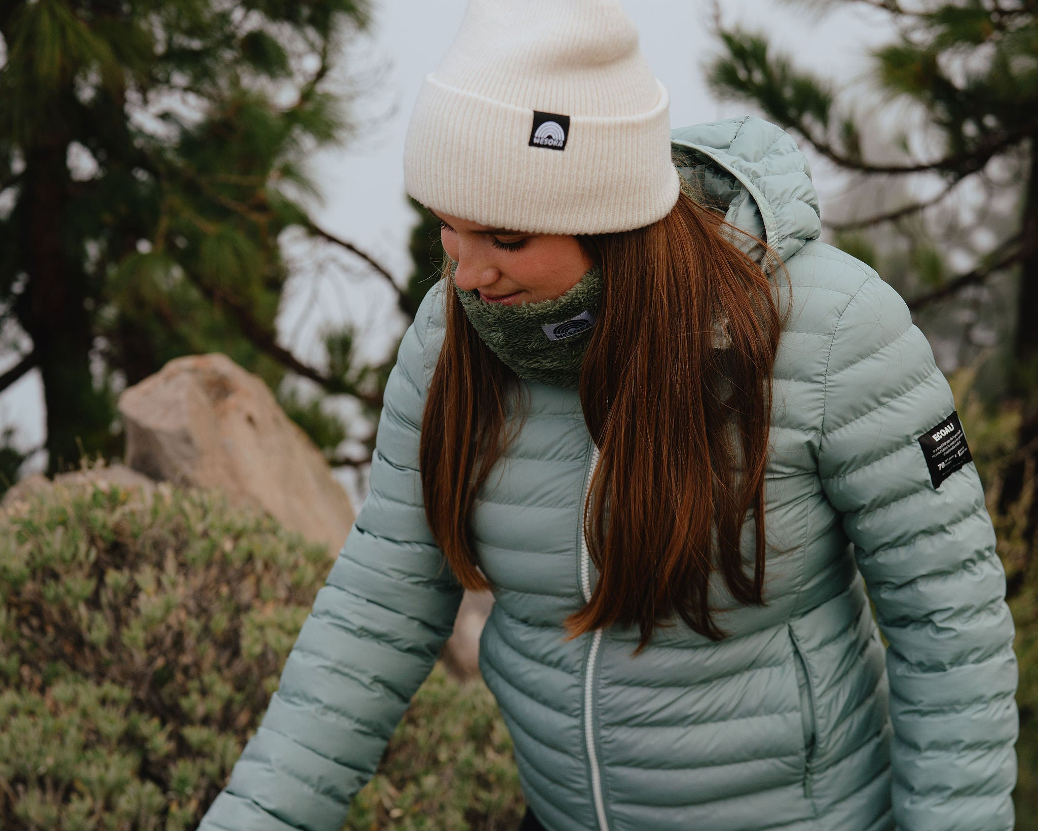Eine Frau läuft durch ein waldähnliches Gebirge. Sie trägt einen weißen Beanie und den Neckwarmer Fluffy Green. Sie ist eins mit der Natur und fässt das Gebüsch an.