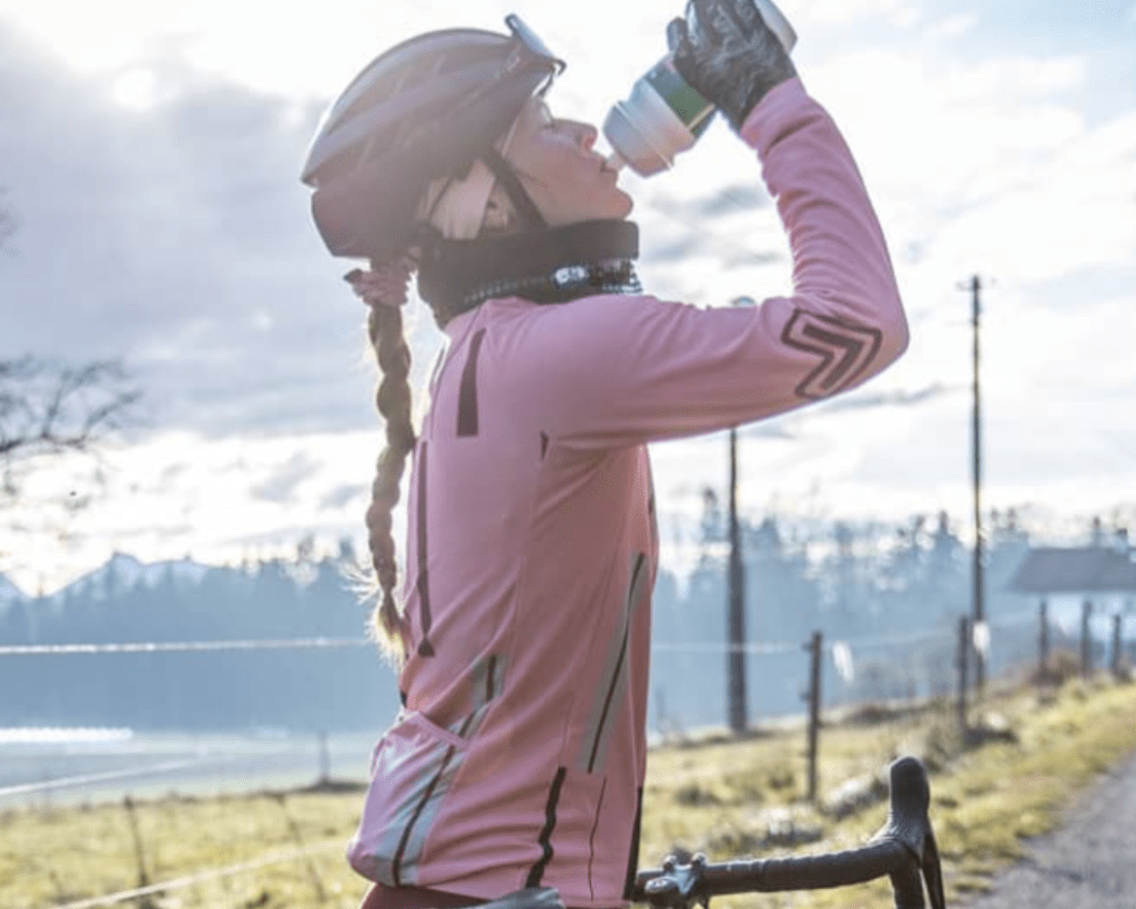 Eine Radfahrerin trägt den Neckwarmer Sparkle Black und ist in vollständiger Radbekleidung. Sie trinkt aus ihrer Flasche. Im Hintergrund sieht man Wälder und Berge