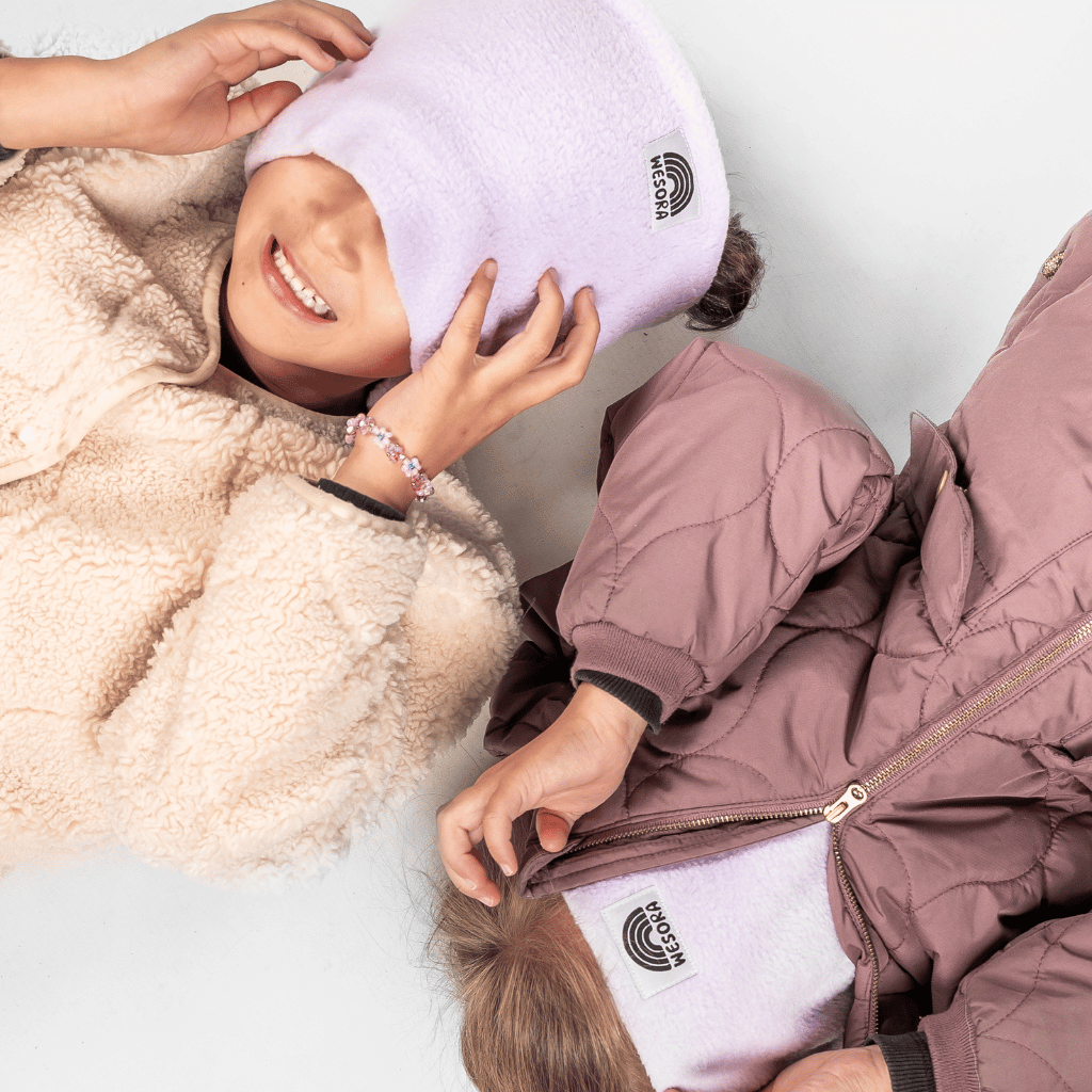 Fleece Neckwarmer - Dreamy Lilac Kid mit kuscheligem Design, getragen von zwei lächelnden Kindern in WESORA-Beanies, liegend auf einer hellen Oberfläche.