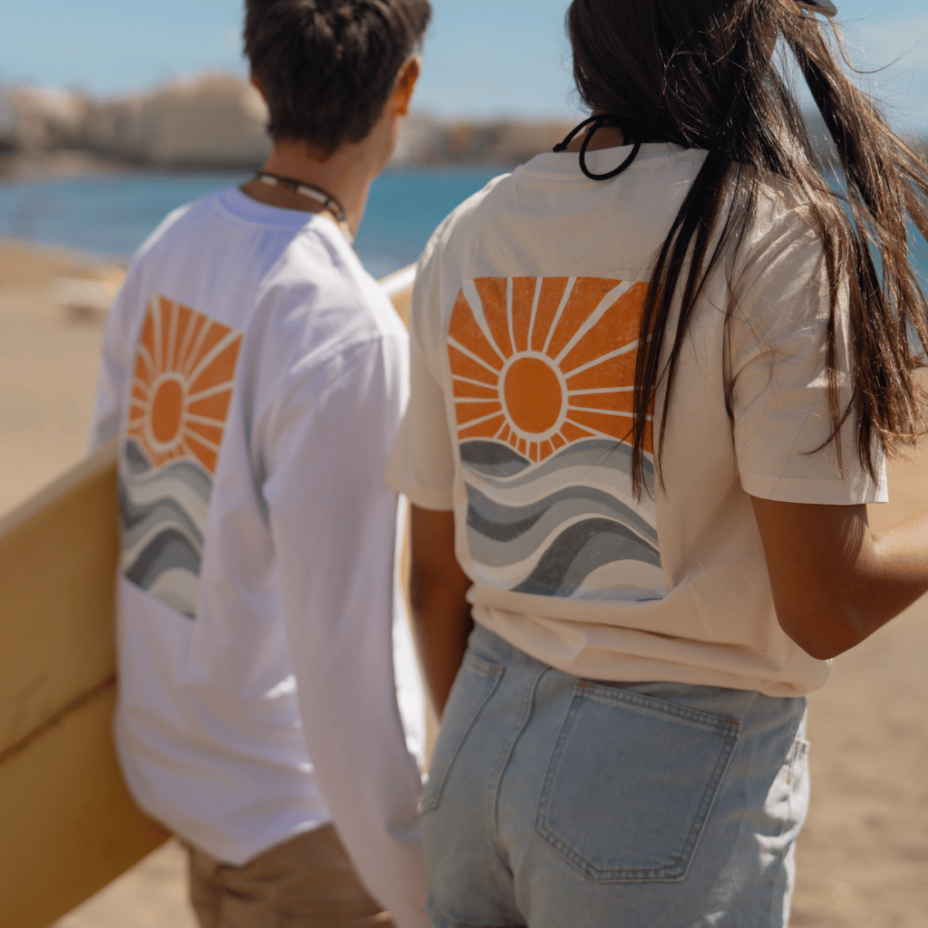 Zwei Personen am Strand mit dem Rücken zur Kamera, tragen Longsleeve Golden Sunrise in weiß mit Sonnen- und Wellenprint; eine Person hält ein Surfbrett, die andere hat lange Haare