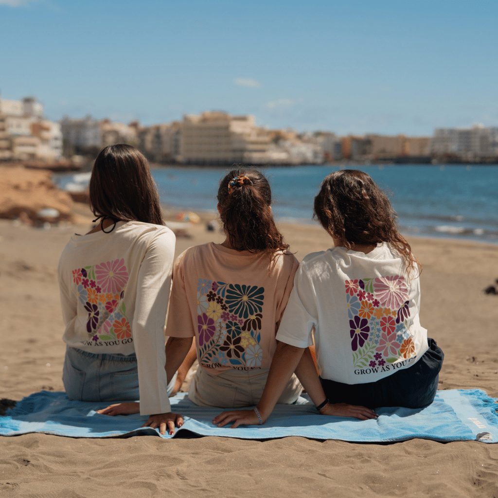 Grow as you go Flower - Longsleeve mit floralem Rückendesign, getragen von drei Frauen sitzend am Strand, eingefangen in entspannter Urlaubsatmosphäre, fotografiert von hinten.