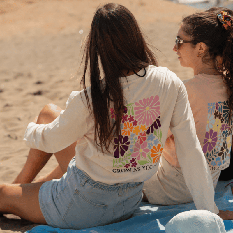 Grow as you go Flower - Longsleeve in weiß getragen von zwei Frauen sitzend am Strand, fotografiert von Hinten mit Blick auf den Druck am Rücken. 