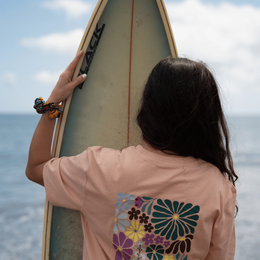 Grow as you go Flower - Unisex Tshirt mit buntem Blumen-Print hinten, von Person mit Surfboard am Strand getragen, Blick aufs Meer.