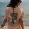 Grow as you go Flower - Unisex Tshirt mit großem Blumenprint und GROW AS YOU GO Schriftzug, getragen von einer Frau am Wasser stehend, Rückenansicht.