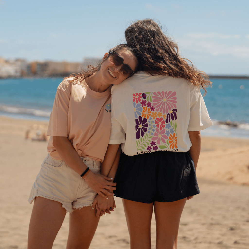 Grow as you go Flower - Unisex T-Shirt mit großem Blumen-Print auf dem Rücken, getragen von zwei Personen am Strand, Hand in Hand, lächelnd.