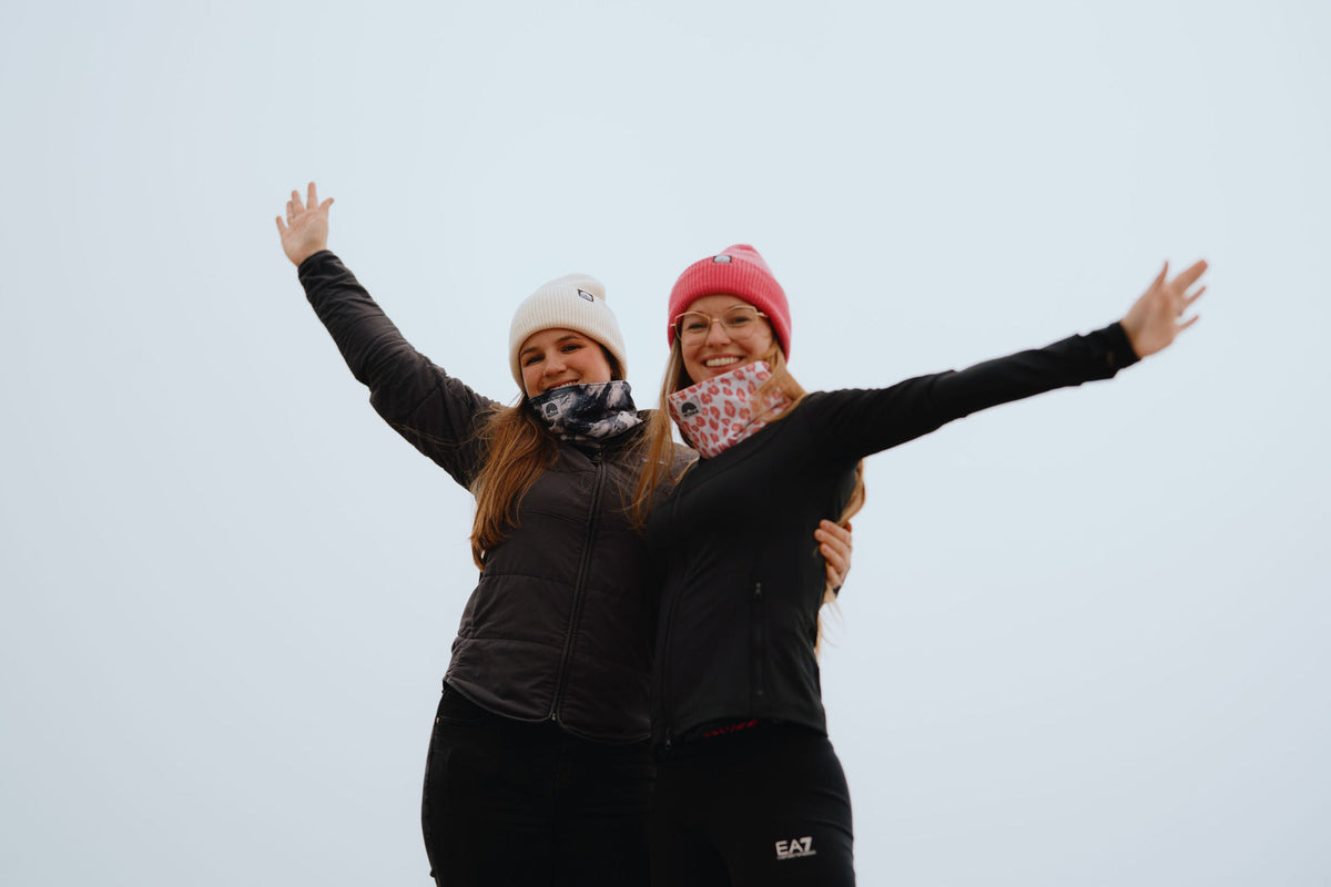 Zwei Frauen mit hochgestreckten Armen tragen Beanie Mützen und Neckwarmer