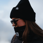 Neckwarmer & Beanie Bundle - Black, getragen von Person mit langem Haar, seitlich fotografiert, in einer kühlen, überbelichteten Umgebung.