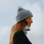 Neckwarmer & Beanie Bundle - Blue, getragen von Person mit rötlich-blondem Haar und klaren Brillen, seitlich vor bewölktem Himmel.