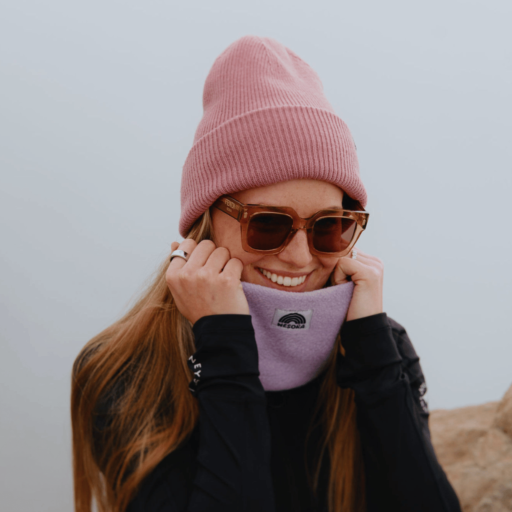 Neckwarmer & Beanie Bundle - Lilac, getragen von einer lächelnden Frau, die den Halswärmer um Kinn und Wangen hält, vor einem neutralen Hintergrund.