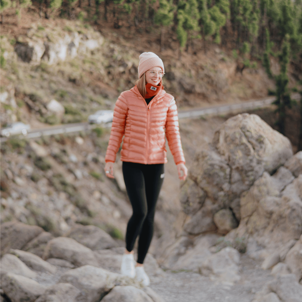 Neckwarmer & Beanie Bundle - Orange, getragen von Person auf felsigem Bergpfad, fotografiert in alpiner Landschaft mit Straßen und Bäumen im Hintergrund.