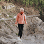 Neckwarmer & Beanie Bundle - Orange, getragen von Person auf felsigem Bergpfad, fotografiert in alpiner Landschaft mit Straßen und Bäumen im Hintergrund.