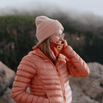 Neckwarmer & Beanie Bundle - Orange, getragen von einer Person in pinker Strickmütze und wattierter Jacke, lächelnd auf einem nebligen Felsvorsprung stehend.