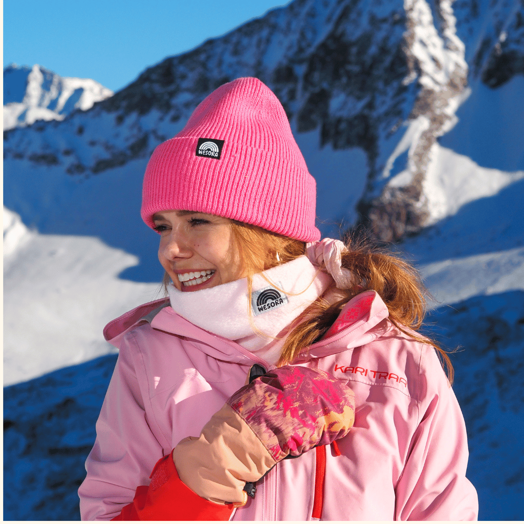Neckwarmer & Beanie Bundle - Rose, getragen von Person in Winteroutfit, lächelnd vor schneebedeckten Bergen, sonniger Tag, Hand am Jackenreißverschluss.