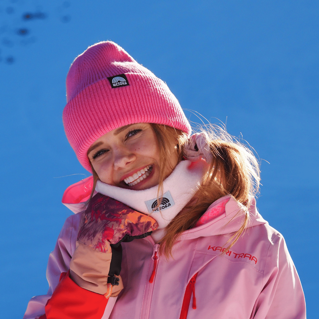 Neckwarmer & Beanie Bundle - Rose, getragen von einer lächelnden Person, in einem Skiresort, Nahaufnahme.