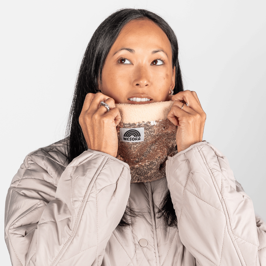 Pailetten Neckwarmer - Sparkle Gold mit roségoldenen Pailletten und weichem Fleece-Innenfutter, gehalten von Frau in beigem Steppmantel.