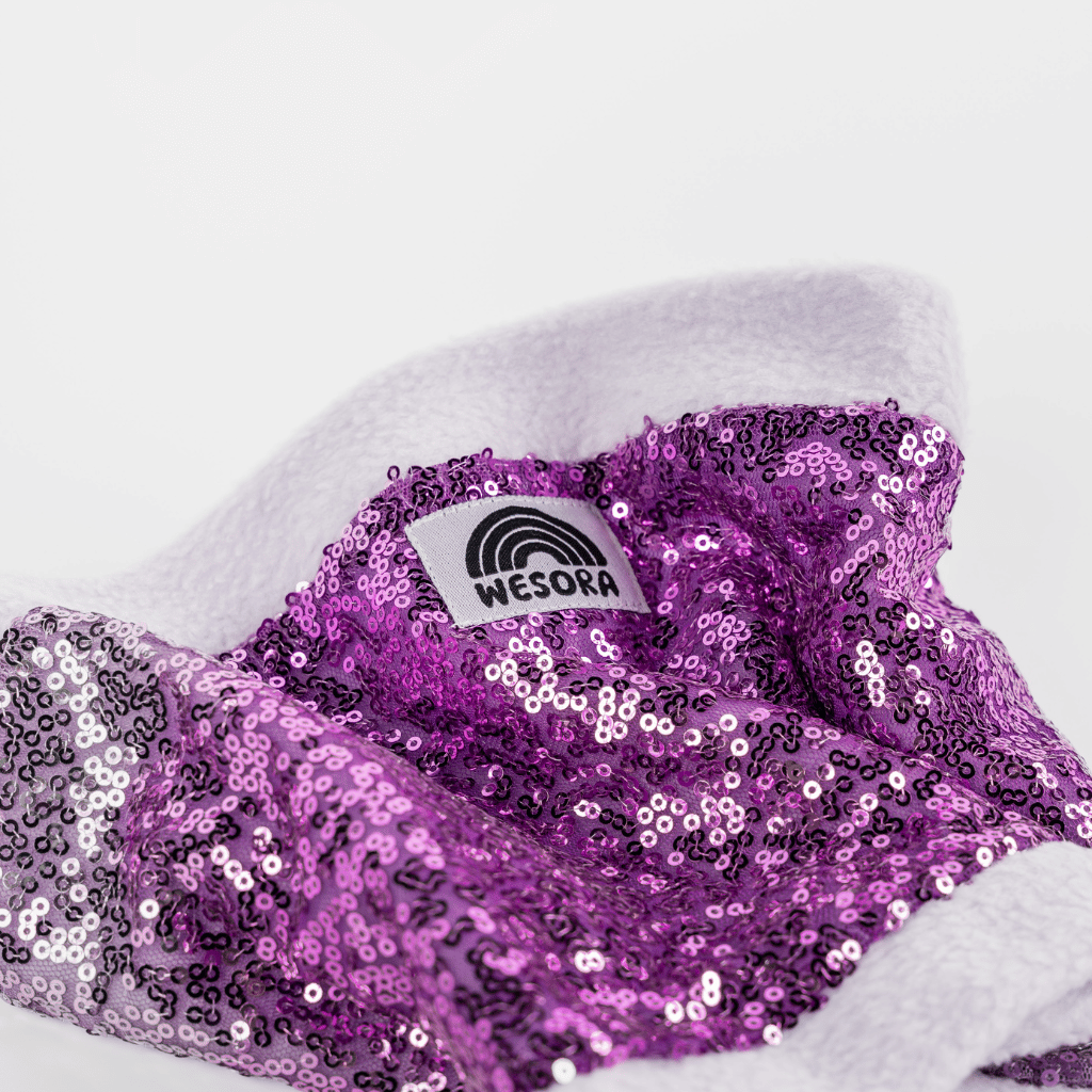 Pailetten Neckwarmer - Sparkle Lilac mit glänzenden lila Pailletten auf weißem Fleece, zeigt WESORA Label, handgemacht und stilvoll für Winterabenteuer.