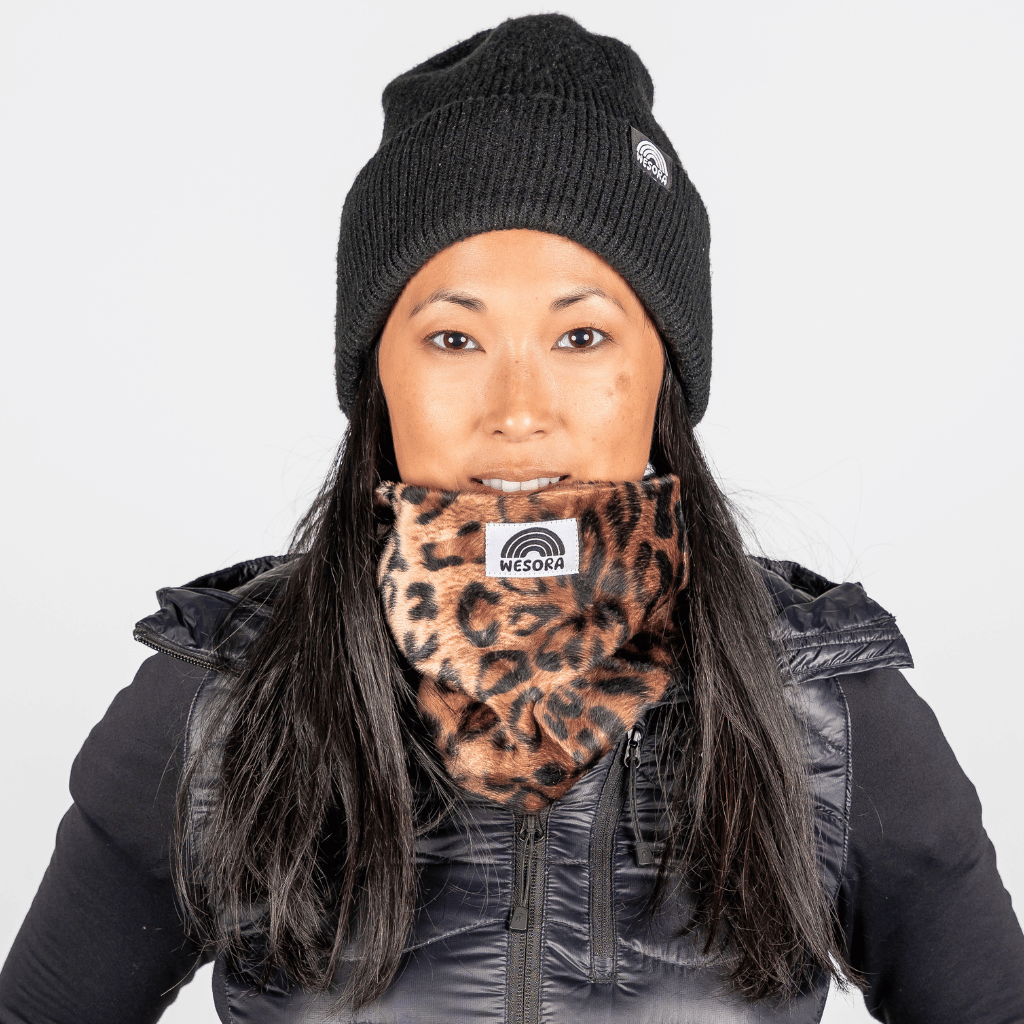 Printed Neckwarmer - Wild Leo Fur mit Leopardenmuster, getragen von Person in schwarzer Weste und Beanie, fotografiert vor neutralem Hintergrund.