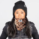Printed Neckwarmer - Wild Leo Fur mit Leopardenmuster, getragen von Person in schwarzer Weste und Beanie, fotografiert vor neutralem Hintergrund.