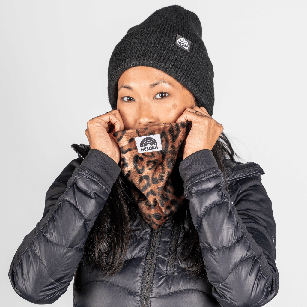 Printed Neckwarmer - Wild Leo Fur mit Leopardenmuster, von Person im schwarzen Outfit vor neutralem Hintergrund präsentiert, zeigt WESORA-Logo.