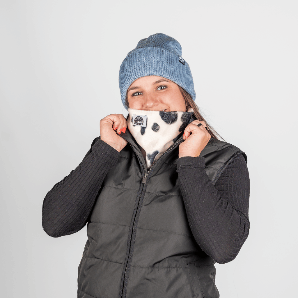 Printed Neckwarmer - Cow Spirit Fur mit Kuhmuster, getragen von Person in Winterkleidung, vor grauem Hintergrund, hält den Neckwarmer vor das Gesicht.